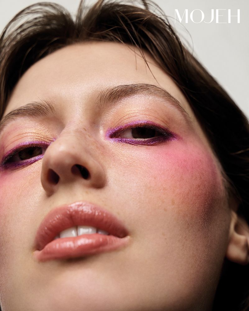 Extreme close-up of a young woman's face with pink and purple makeup, and moist lips, set against a stark white background.