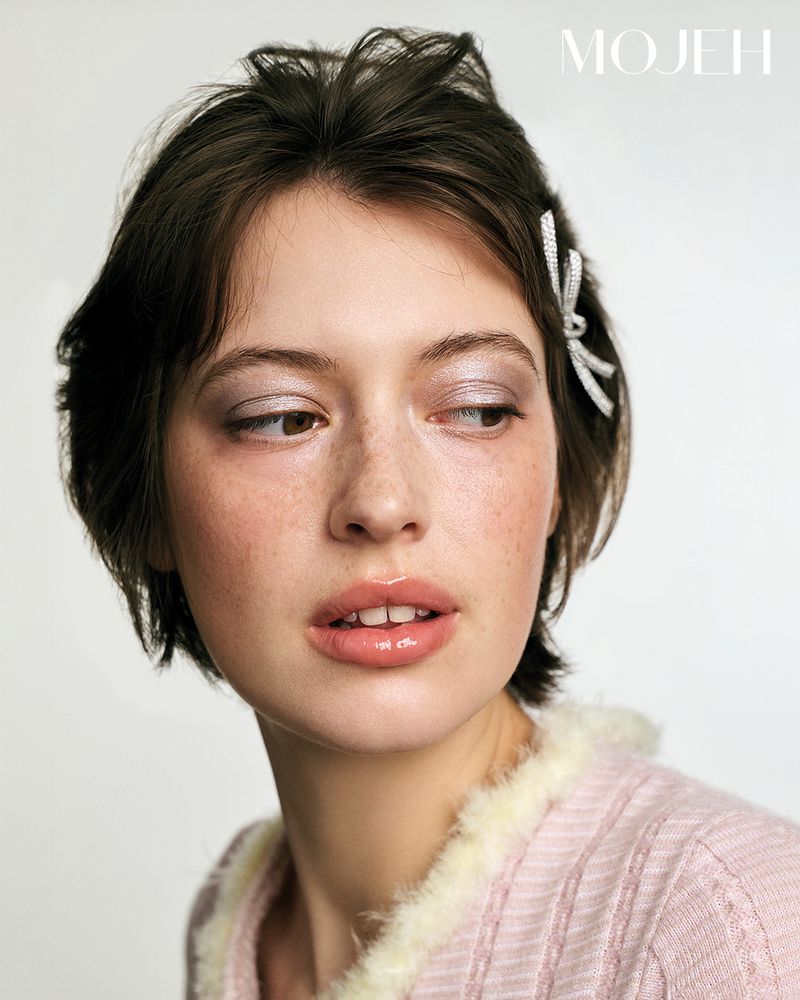 Close-up portrait of a young woman with freckles, wearing a pink sweater and hair clip, against a white backdrop. Beauty shot.