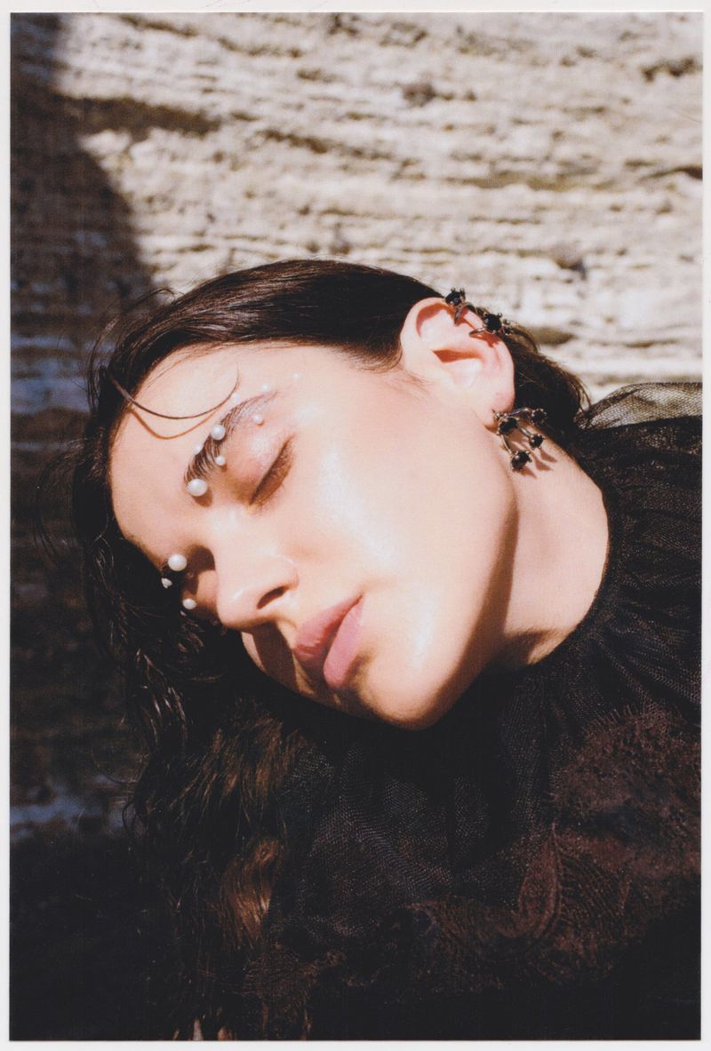 Portrait of a young woman with closed eyes wearing black lace and pearls, against a textured light brown and tan stone wall.