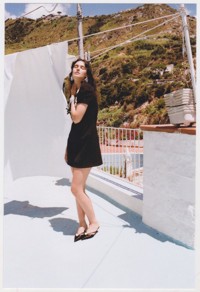 A young woman in a black dress stands on a rooftop with a mountain view, posing near a white sheet on a sunny day.