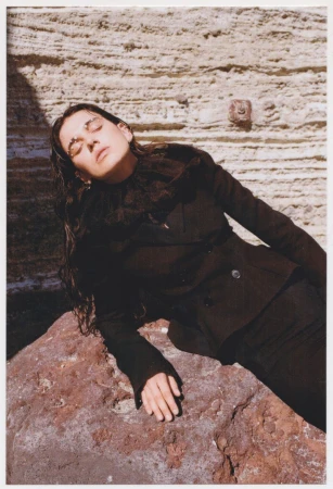 Fashion shot of a brunette woman in a black jacket and skirt, reclining on a red rock with a textured rock wall backdrop in natural light.