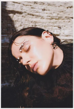 Close-up portrait of a woman with pearls on her eyebrows wearing a dark ruffled top against a textured stone wall background