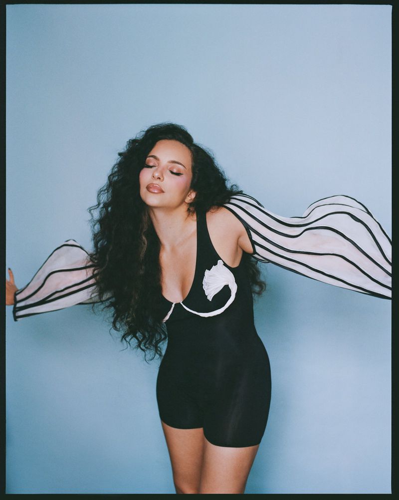 Portrait of a woman with long curly hair, wearing a black bodysuit with white accents, posing against a light blue background.