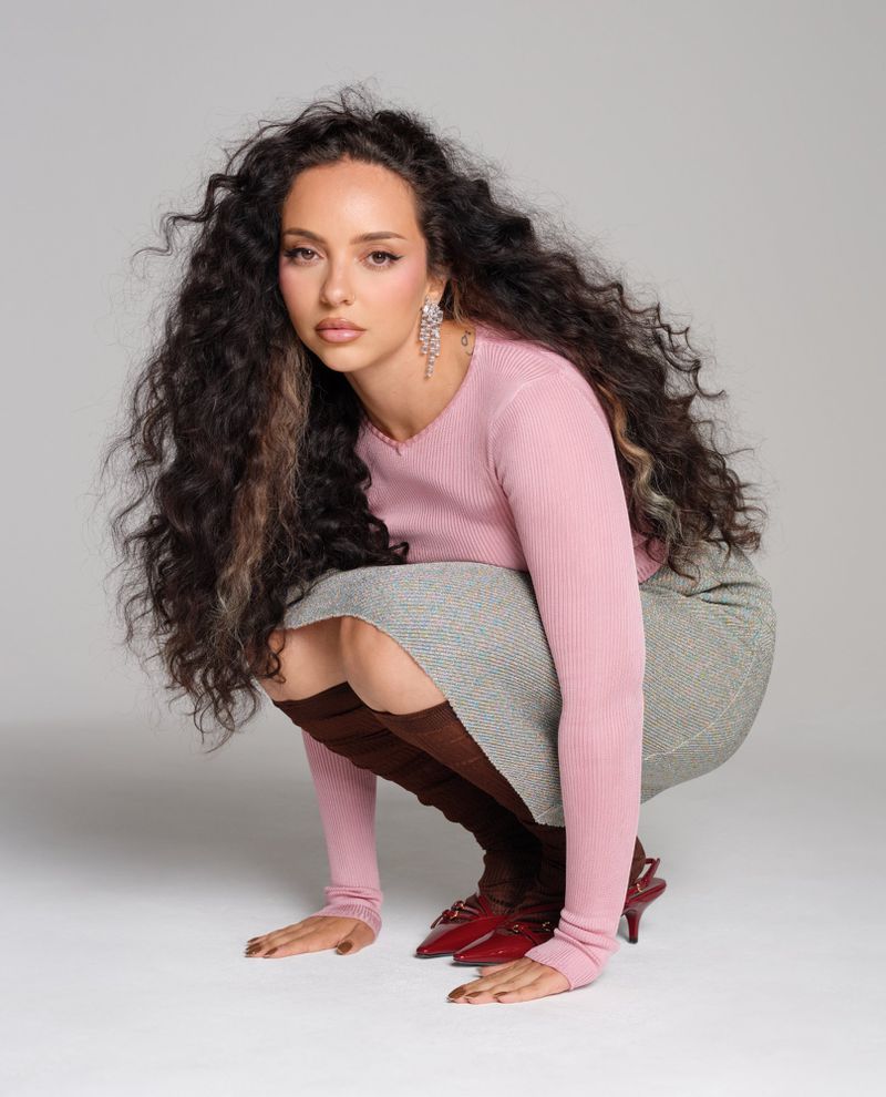 Young woman with curly hair poses in a pink sweater and grey skirt in a photo studio setting against a plain backdrop.