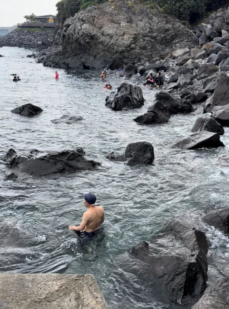 A person sits in the shallow ocean water, surrounded by dark rocks, on a cloudy day; other swimmers are also visible in the background.