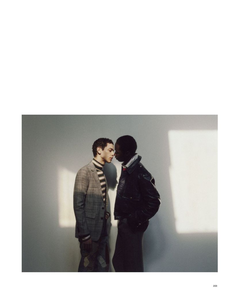 Two men stand indoors, facing each other closely. One man wears a grey jacket and striped turtleneck. The other wears a black leather jacket.