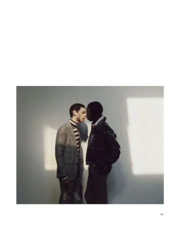 Two men stand indoors, facing each other closely. One man wears a grey jacket and striped turtleneck. The other wears a black leather jacket.