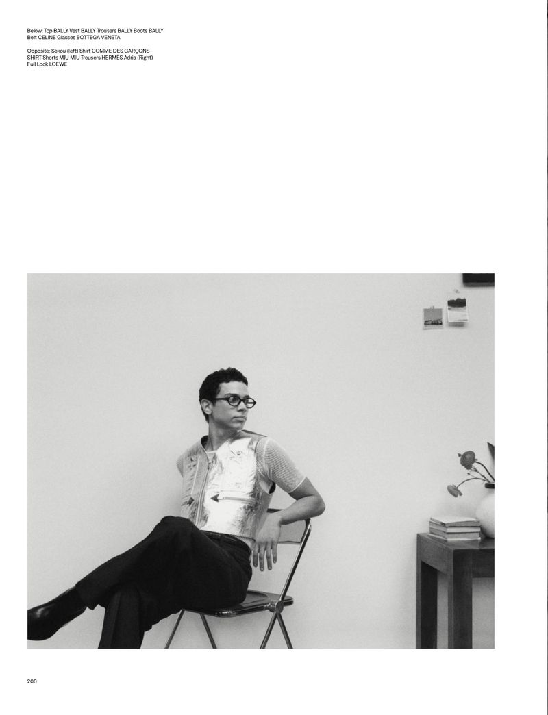 Man in glasses sits on a folding chair in a photo studio wearing a sheer top and vest, flowers and books on a nearby table. 