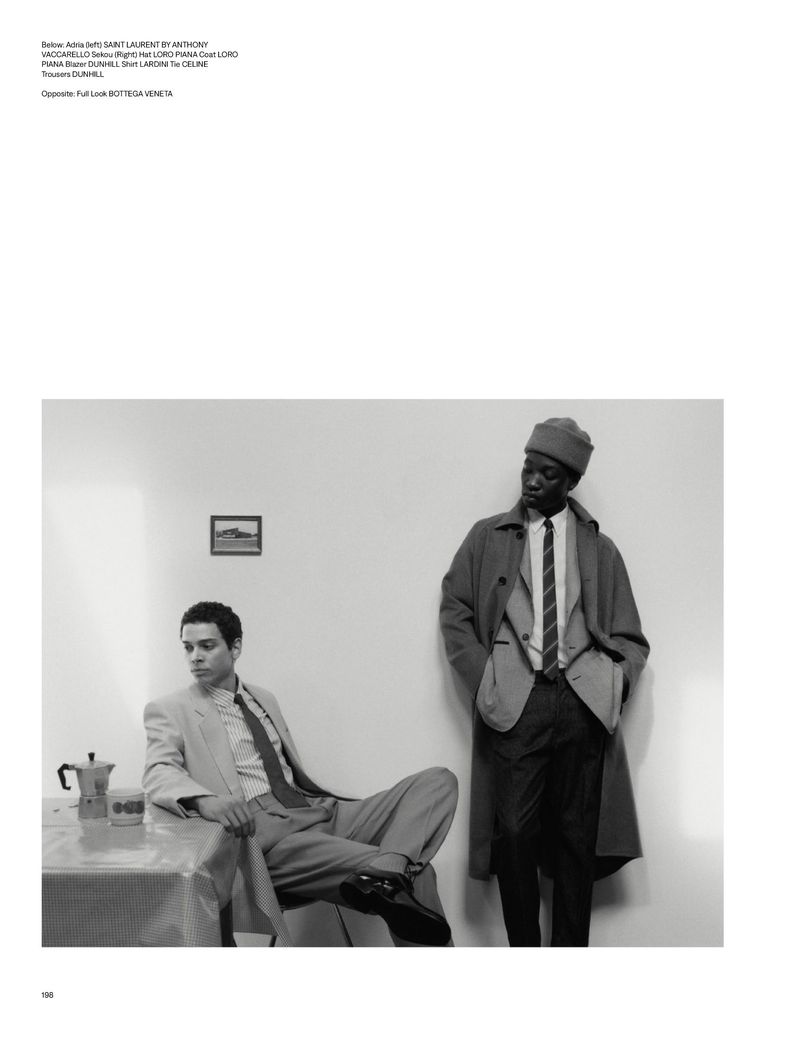 Two men in retro-style suits in a minimal indoor setting. One seated by a table with a coffee pot, the other standing.
