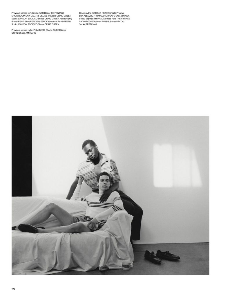 Monochrome fashion shot: Two young adult men in casual wear pose on a white couch with shoes nearby against a neutral wall.