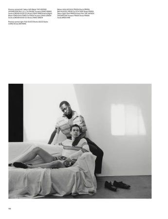 Monochrome fashion shot: Two young adult men in casual wear pose on a white couch with shoes nearby against a neutral wall.