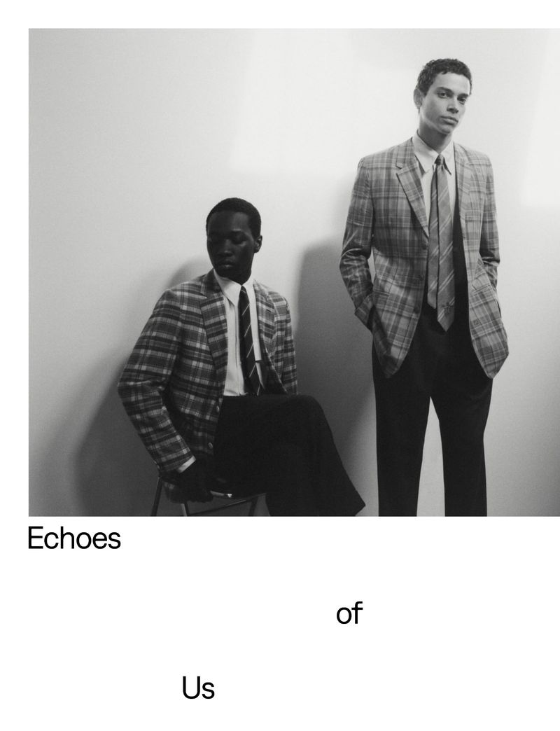 Two men in plaid blazers pose in a black and white fashion shot. One man sits while the other stands. Studio lighting highlights the texture.
