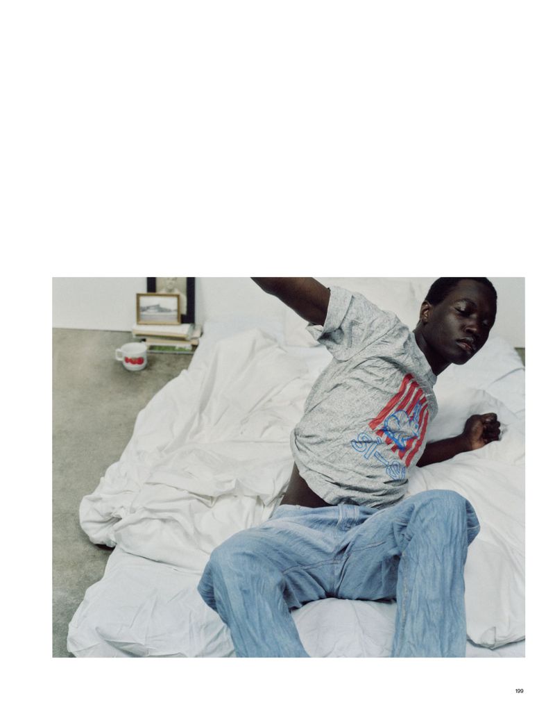 A young adult male lounges on a white bed with a grey patterned t-shirt and blue jeans in an indoor setting with natural lighting.