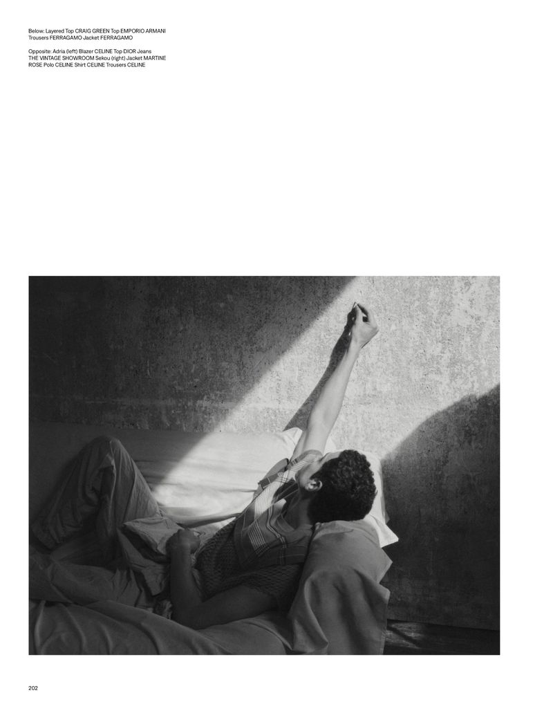 Monochromatic portrait of a man lying on a bed, arm raised toward a light beam, in a room with a textured wall, fashion editorial.
