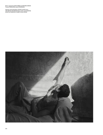Monochromatic portrait of a man lying on a bed, arm raised toward a light beam, in a room with a textured wall, fashion editorial.