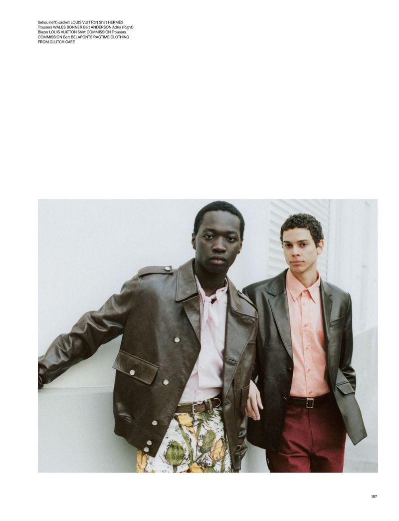 Two male models in leather jackets and patterned pants pose indoors against a neutral wall, showcasing high-end fashion.