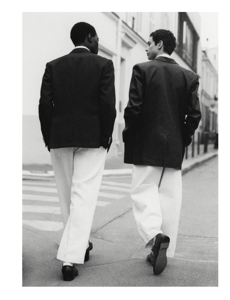 Two adult men in suits and pants walk away from the camera on a city street with buildings and a crosswalk. Black and white.