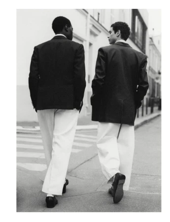 Two adult men in suits and pants walk away from the camera on a city street with buildings and a crosswalk. Black and white.