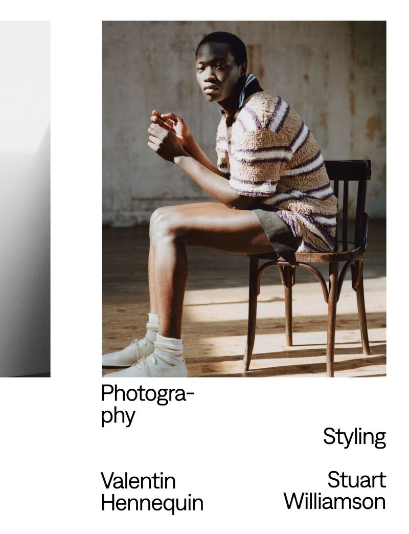 A young adult sits on a wooden chair, inside with natural light, modeling a striped top and shorts, wearing white sneakers and socks.