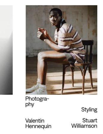 A young adult sits on a wooden chair, inside with natural light, modeling a striped top and shorts, wearing white sneakers and socks.