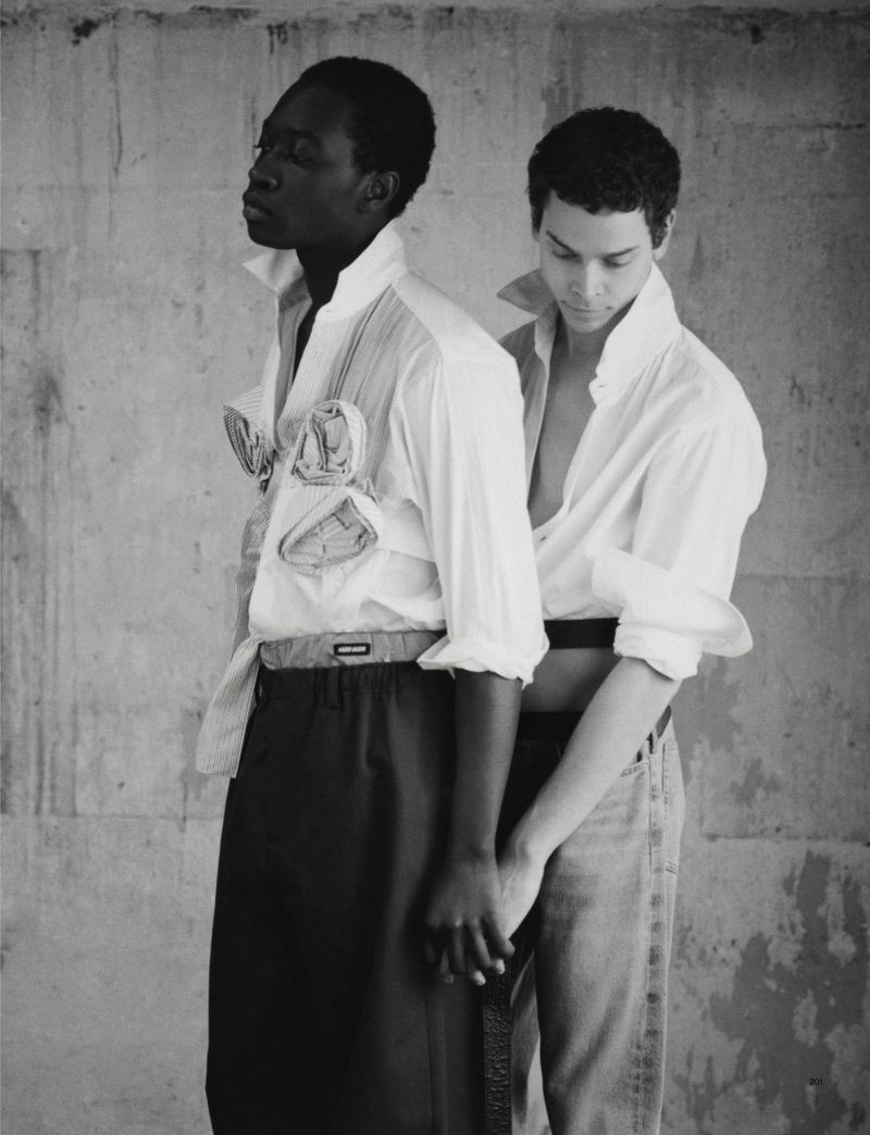 Monochrome fashion portrait of two men in unbuttoned white shirts against a textured wall, holding hands in a studio environment.