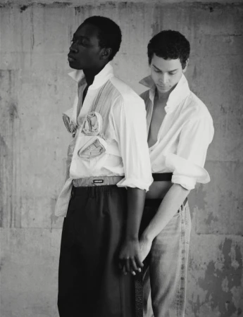 Monochrome fashion portrait of two men in unbuttoned white shirts against a textured wall, holding hands in a studio environment.