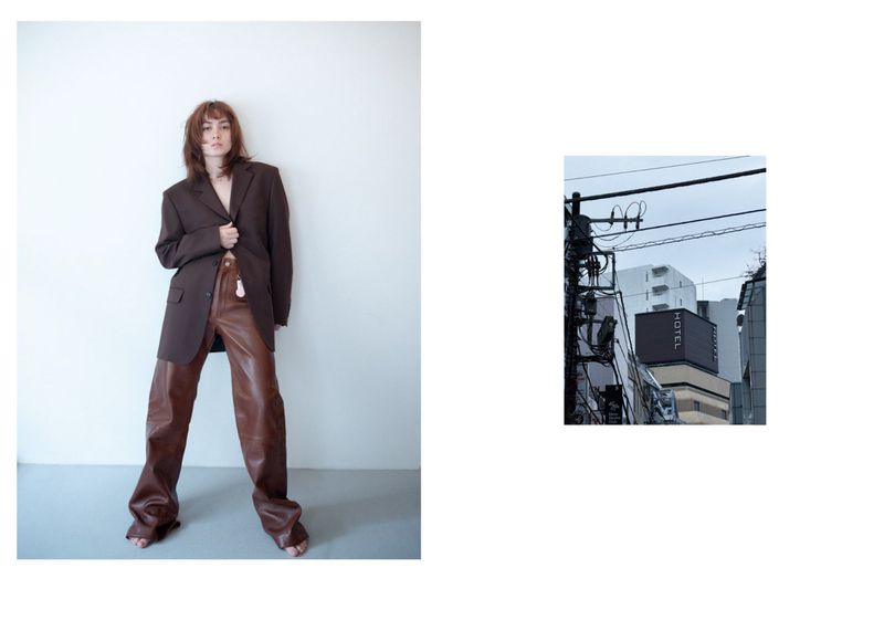 A full-length fashion shot of a woman in an oversized brown blazer and leather pants against a white wall in a photo studio.