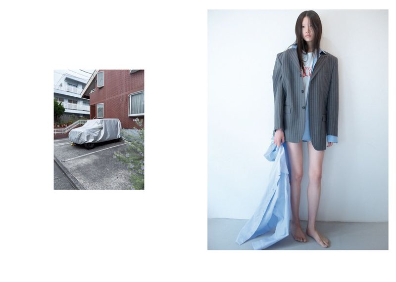Fashion shot of a slender young woman in an oversized pinstripe blazer, holding a light-blue shirt, standing against a white wall; car in background.