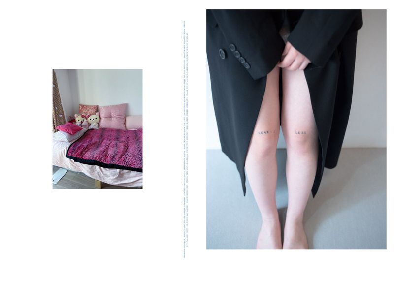 An individual in a black coat stands against a grey wall with bare legs that feature the tattoos 'love' and 'less'.
