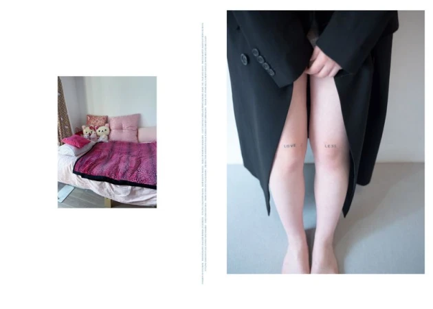 An individual in a black coat stands against a grey wall with bare legs that feature the tattoos 'love' and 'less'.