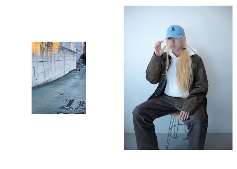 Person in stylish streetwear sits on a stool indoors in front of a white wall wearing a hoodie, jacket, hat, and brown pants.