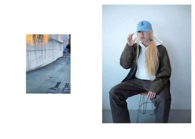 Person in stylish streetwear sits on a stool indoors in front of a white wall wearing a hoodie, jacket, hat, and brown pants.