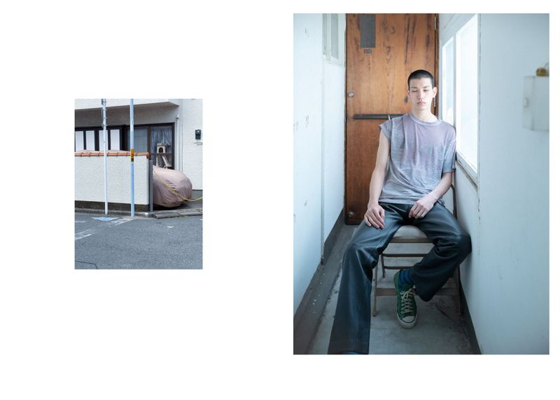 A slender, neutral-expression young man sits on a metal chair near a window in an indoor setting with walls, wearing contemporary clothing.