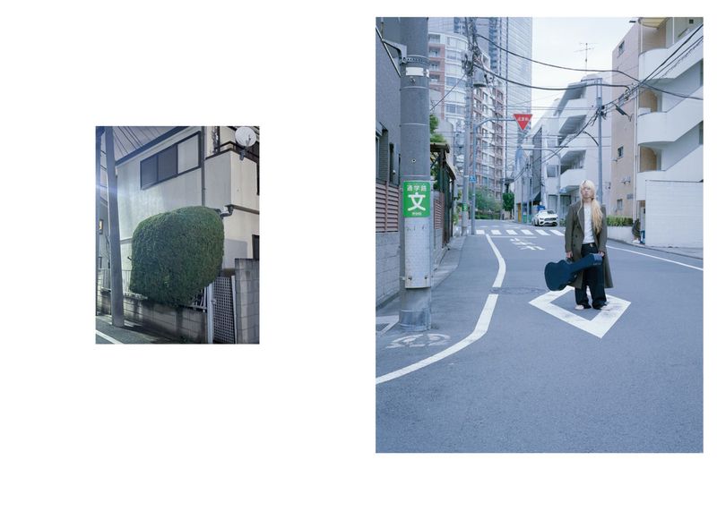 A person with long blond hair holding a guitar case stands on a crosswalk in a street in Japan with buildings in the background.