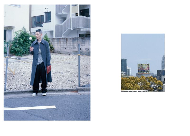 An Asian man in a trench coat holding a baseball in an urban setting with an apartment building and distant city skyline visible.