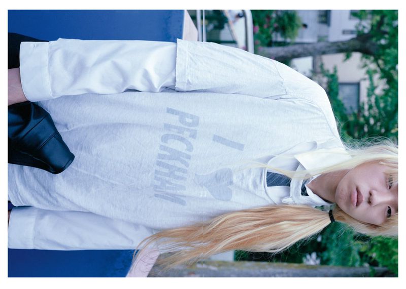 Teen fashion: Person with blond ponytail wears a white shirt that says 'I heart Peckham,' set against a backdrop of green foliage.