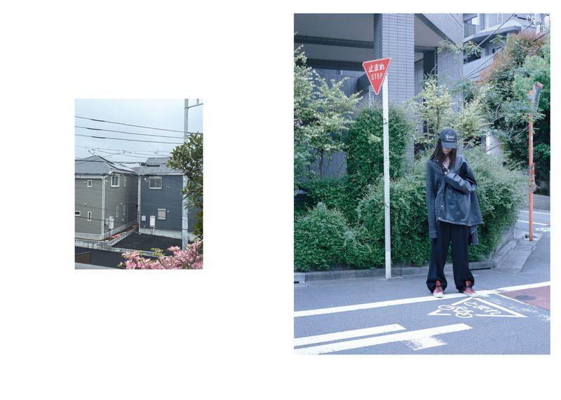 An adult female stands on a street next to a stop sign and green foliage, wearing a dark outfit and cap, in a daylight outdoor scene.