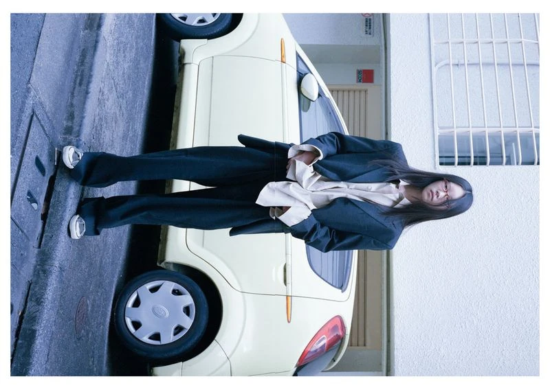 Stylish woman poses in front of a light-yellow car on a city street, wearing a blazer, trousers, and sneakers in an urban environment.