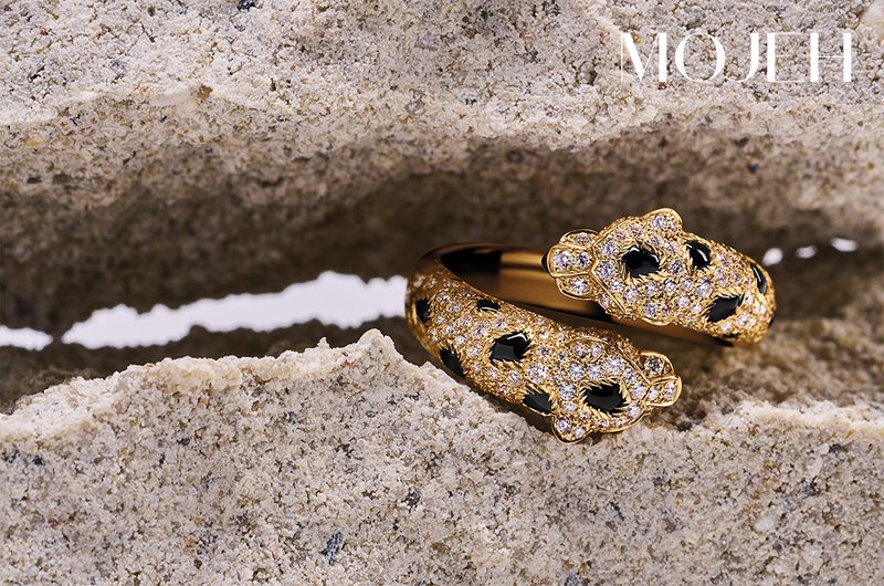Gold ring featuring diamonds and black gems in an animalistic style. The ring is displayed on a textured, sandy surface.