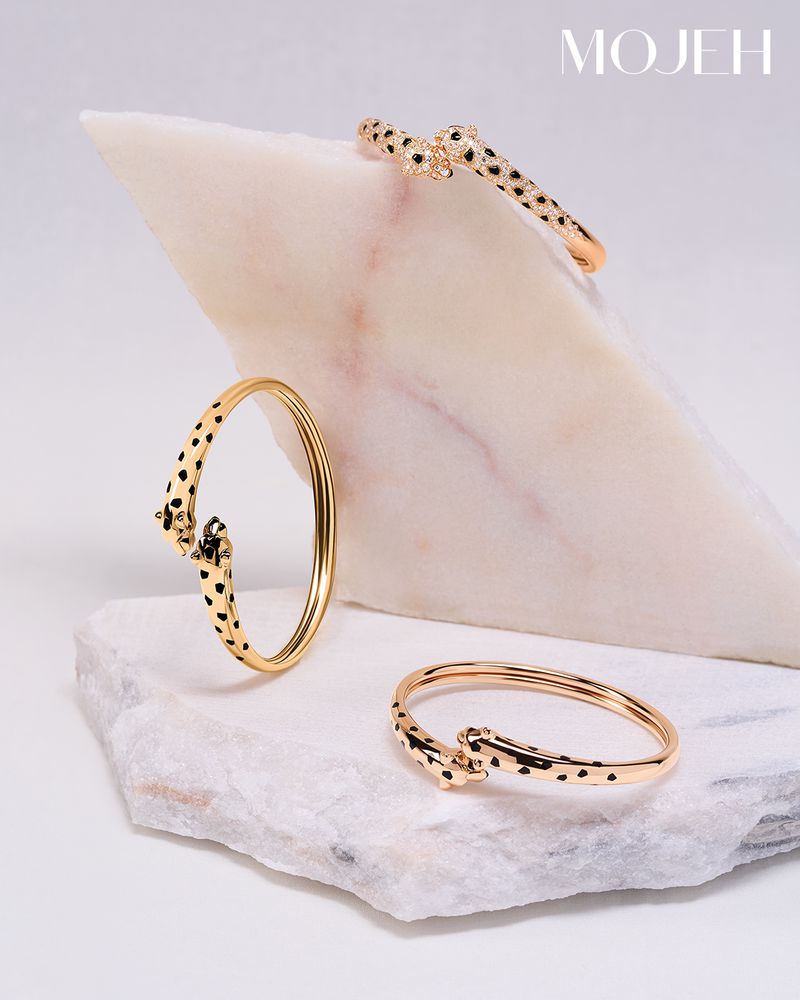Three gold-toned bracelets with black cheetah spots are styled on white marble, highlighted by studio lighting for jewelry advertisement.
