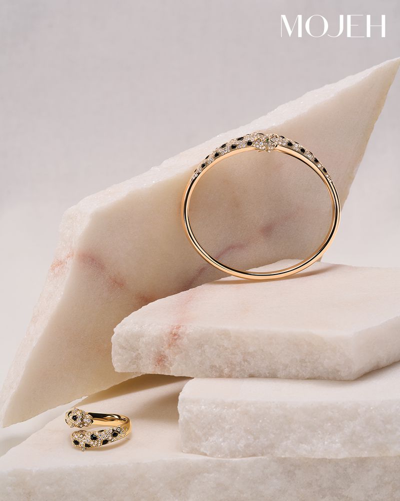 Still life of gold jewelry, including a bracelet and ring, with black and white accents, displayed on marble slabs against a pale background.