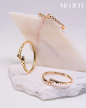 Three gold leopard motif cuff bangles arranged on angular white and pale pink marble slabs against a plain studio background.