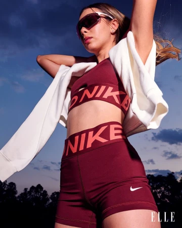 Woman in Nike sportswear poses outdoors against a blue sky, wearing sunglasses and a jacket draped over her shoulders for a sporty look.