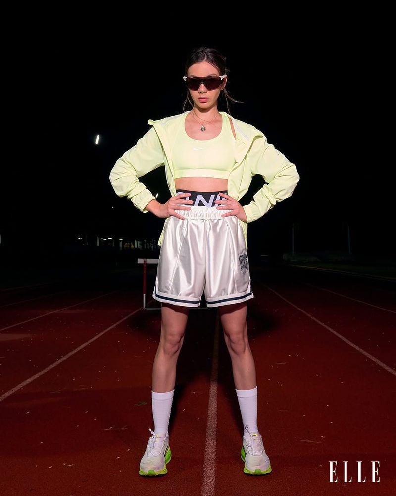 A woman in athletic wear stands on a running track at night, wearing sunglasses, a neon yellow sports bra, matching jacket, and white athletic shorts.