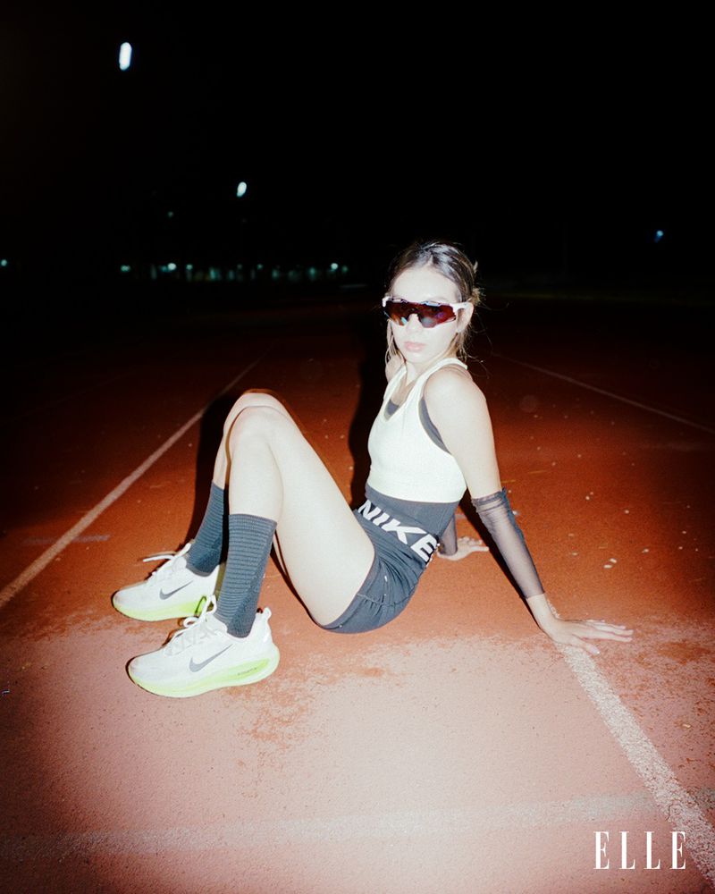 Female athlete sitting on a red track with Nike athletic wear, sunglasses, and running shoes, with an outdoor running track setting.
