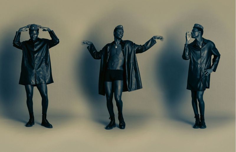 Three identical African American adult males pose in a studio wearing black coats, shorts, caps, and boots against a neutral background.