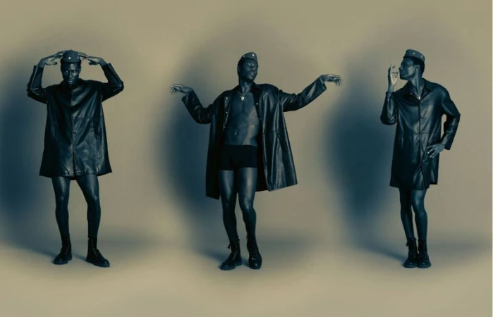 Three identical African American adult males pose in a studio wearing black coats, shorts, caps, and boots against a neutral background.