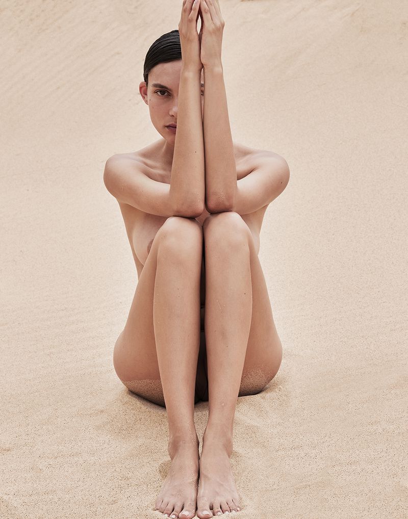 Topless woman with arms raised and hands clasped sits in sand, eyes forward, in an outdoor tan sand setting in soft, diffused light.