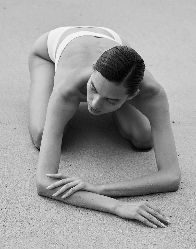 Monochrome portrait of an adult woman kneeling on a sandy surface, wearing a swimsuit, with hands crossed on the ground.