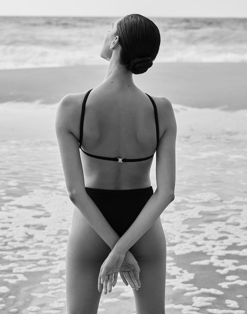 Back view of a woman in a black bikini standing in the ocean with her hands crossed behind her back on a sandy beach.